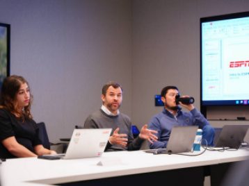 ESPN employee gesticulates while giving a presentation, with two other employees sitting at their side.